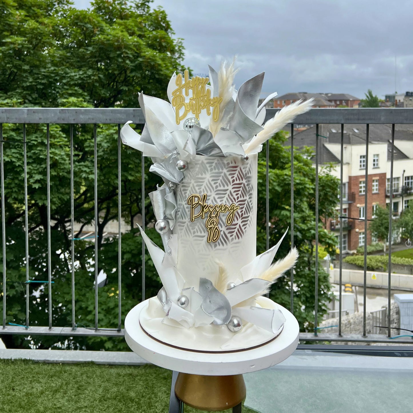 Decorative cake with white and gold wafer paper elements on a balcony with greenery and buildings in the background
