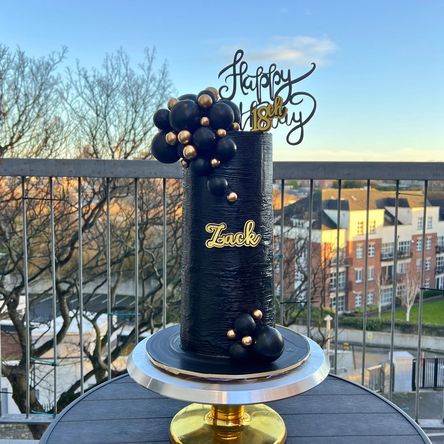 Black birthday cake with gold accents on a balcony with a cityscape background