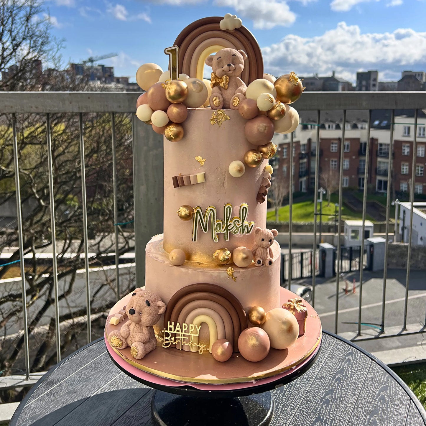 Decorative cake with teddy bears, rainbow, and balloons on a rooftop with cityscape background