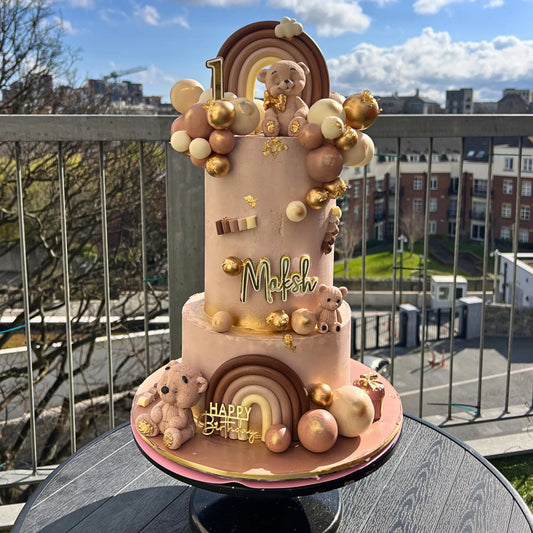 Decorative cake with teddy bears, rainbow, and balloons on a rooftop with cityscape background