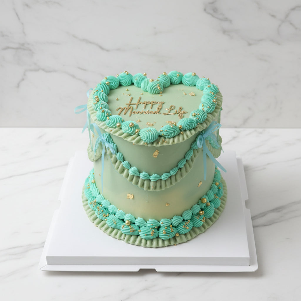 A two-tiered round cake decorated with mint green frosting, piped turquoise rosettes, gold leaf accents, and 'Happy Married Life' written in gold icing on top, displayed on a white box outdoors.