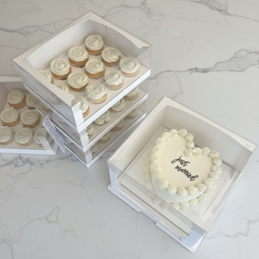 White bakery boxes containing frosted cupcakes and a heart-shaped cake with piped cream borders and the words 'just married' in script, arranged on a tabletop.